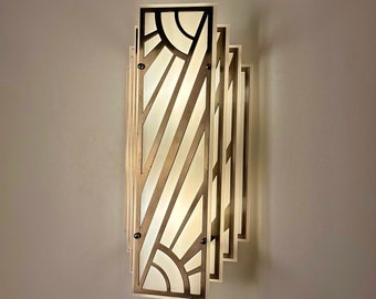 May include: A gold-toned wall sconce with a geometric design. The sconce has a white frosted glass panel and a layered design with multiple panels.