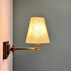 K&ouml;nnte beinhalten: Eine Wandleuchte aus Messing mit beigefarbenem Stoffschirm. Die Lampe hat einen verstellbaren Arm und einen rechteckigen Sockel. Der Schirm ist konisch geformt und die Lampe ist an einer wei&szlig;en Wand mit tealfarbenem Hintergrund befestigt.
