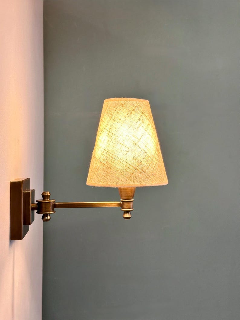 K&ouml;nnte beinhalten: Eine Wandleuchte aus Messing mit beigefarbenem Stoffschirm. Die Lampe hat einen verstellbaren Arm und einen rechteckigen Sockel. Der Schirm ist konisch geformt und die Lampe ist an einer wei&szlig;en Wand mit tealfarbenem Hintergrund befestigt.