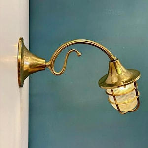 Könnte beinhalten: Eine Wandleuchte aus Messing mit geschwungenem Arm und einem Lampenschirm aus Glas mit Käfig. Die Leuchte ist an einer weißen Wand mit türkisfarbenem Hintergrund befestigt. Das Licht ist an.