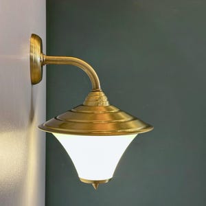 Puede incluir: Una lámpara de pared de latón con una pantalla de vidrio blanco. La lámpara tiene un brazo curvo y una base redonda.