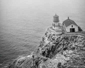 May include: A black and white photograph of a lighthouse on a cliff overlooking the ocean. The lighthouse is a white building with a black roof and a black lantern on top. The ocean is calm and the sky is cloudy.