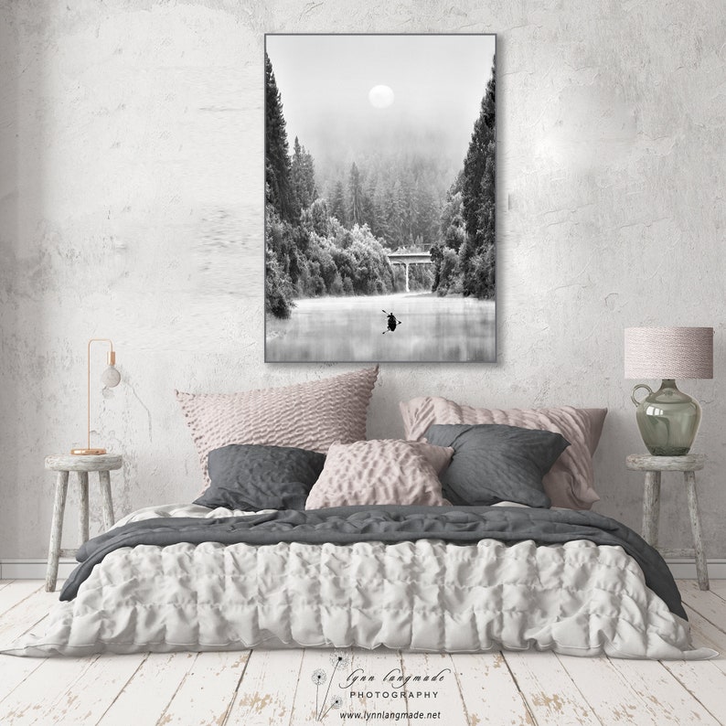 May include: Black and white framed photograph of a misty river scene with a bridge and a person in a kayak. The photograph is hanging above a bed with decorative pillows and a white comforter. The photograph is labeled "Lynn Langmade PHOTOGRAPHY".
