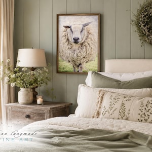 May include: A framed wall art print featuring a front-facing, thick-fleeced sheep standing in a green field. The artwork is displayed in a wooden frame on a sage green paneled wall above a bed with neutral-toned bedding and decorative pillows.