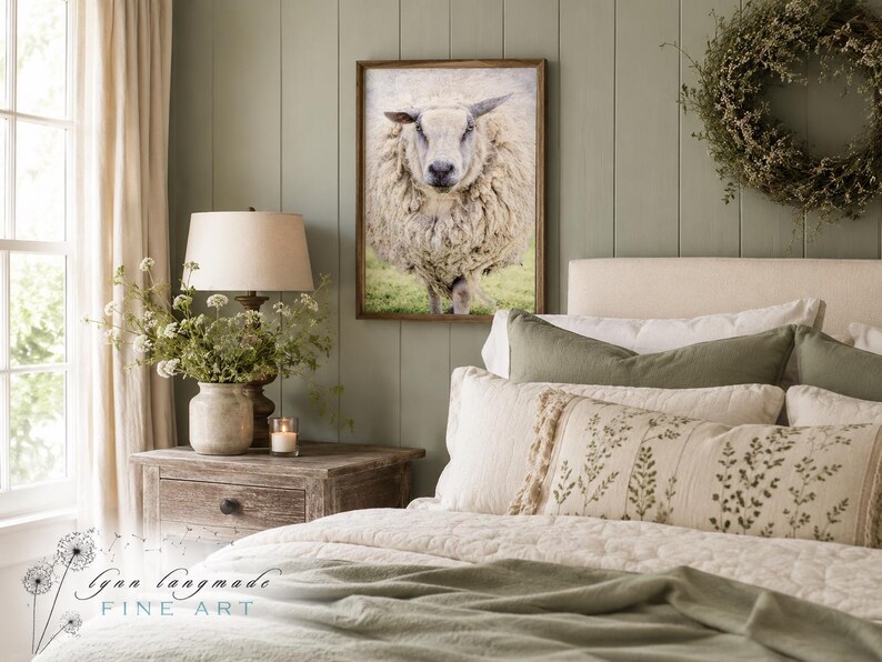 May include: A framed wall art print featuring a front-facing, thick-fleeced sheep standing in a green field. The artwork is displayed in a wooden frame on a sage green paneled wall above a bed with neutral-toned bedding and decorative pillows.