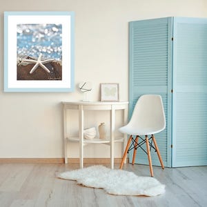 May include: A framed photograph featuring a white starfish on a weathered surface, set against a blurred blue and white background. The photograph is in a light blue frame, displayed on a white wall. The room includes a white table, chair, and a light blue folding screen.