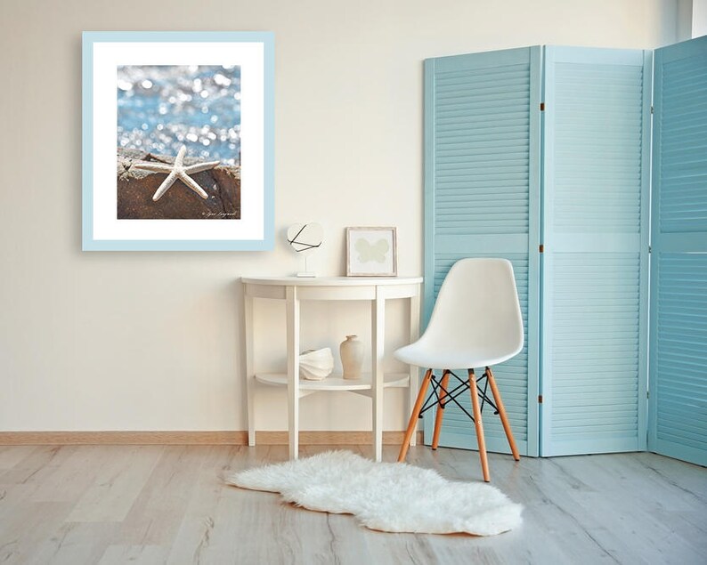 May include: A framed photograph featuring a white starfish on a weathered surface, set against a blurred blue and white background. The photograph is in a light blue frame, displayed on a white wall. The room includes a white table, chair, and a light blue folding screen.