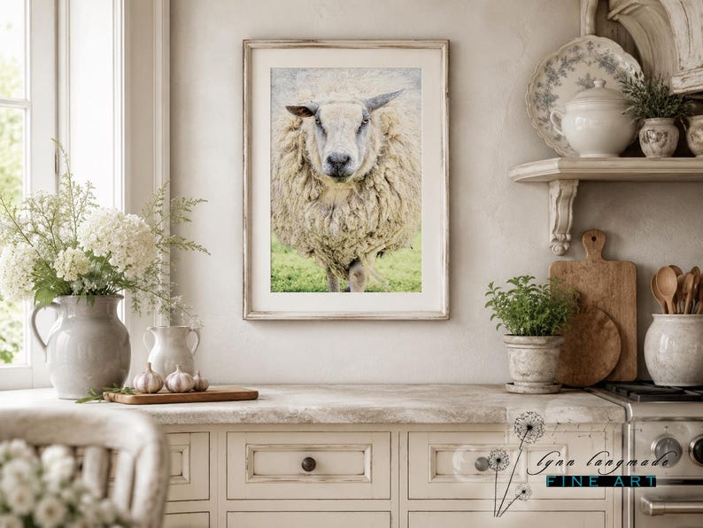 May include: A framed wall art print featuring a front-facing, thick-fleeced sheep standing in a green pasture. The artwork is displayed in a light-colored wooden frame, showcasing the detailed texture of the sheep's wool and its calm expression.