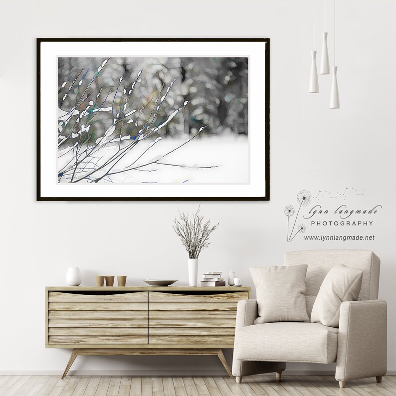 Op de afbeelding: Een ingelijste kunstdruk met een winterlandschap met met sneeuw bedekte takken op de voorgrond. Het kunstwerk toont een vage achtergrond van bomen en sneeuw. De print wordt weergegeven boven een houten kast en een beige fauteuil.