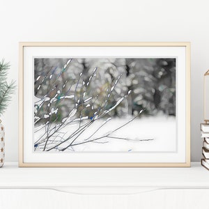 Op de afbeelding: Een ingelijste kunstdruk met een winterlandschap met besneeuwde takken. Het kunstwerk zit in een gouden lijst en wordt op een wit oppervlak weergegeven. Een kleine kerstboom in een decoratieve pot en een kaars in een gouden lantaarn staan ook op het oppervlak.