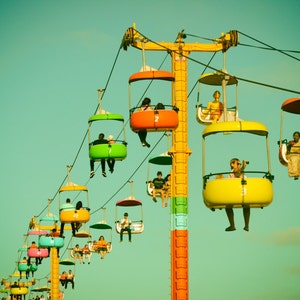 May include: A vintage-toned image of a colorful amusement park ride. Several gondolas in shades of green, orange, yellow, and pink are suspended from a tall, yellow support structure. People are riding in the gondolas against a teal sky.