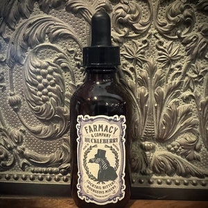 May include: A brown glass bottle with a dropper top and a label that reads "Farmacy & Company Huckleberry Made in Montana" with a black and white zebra illustration. The label also says "Cocktail Bitters Bourgeois Mixers Premium Blend".