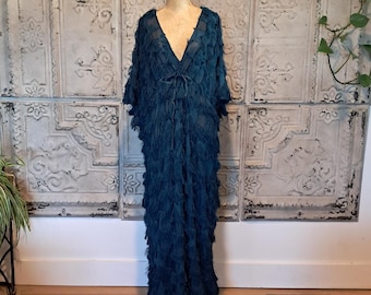 May include: A floor-length, deep teal kaftan dress featuring a V-neckline, a drawstring waist tie, and wide sleeves. The entire garment is covered in a dense, layered fringe texture that creates a voluminous, flowing silhouette.