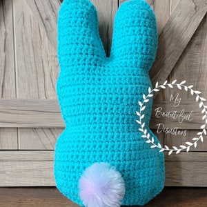 May include: A turquoise blue crocheted bunny cushion with a white and pink fluffy tail. The cushion is sitting on a wooden surface.
