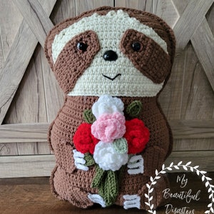 May include: A crocheted sloth holding a bouquet of red, pink, and white flowers. The sloth is brown and white with black eyes. The flowers are surrounded by green leaves. The sloth is sitting on a wooden surface. The text "My Beautiful Disasters" is written in a white wreath on the right side of the image.