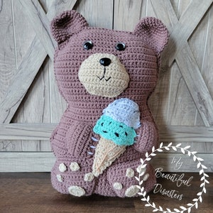 May include: A brown crocheted teddy bear holding a crocheted ice cream cone with blue and green ice cream. The bear has black eyes and a brown nose. The background is a wooden wall with a white X pattern.