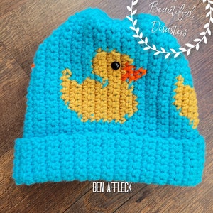 May include: A blue crocheted hat with a yellow rubber ducky design. The hat has a fold-over brim and the text "BEN AFFLECK" is written on the brim.
