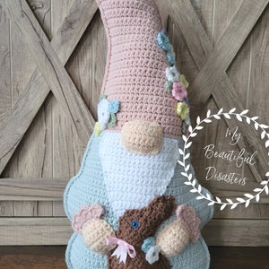 May include: A crocheted gnome doll with a pink hat, white beard, and blue body. The gnome is holding a brown crocheted bunny with a pink bow. The gnome is decorated with pastel colored flowers.