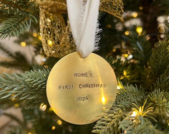 以下が含まれることがあります： 「ROME'S FIRST CHRISTMAS 2024」と刻まれた、丸い金色のクリスマスオーナメント。白いリボンで吊るされており、金色のリボンとライトで飾られたクリスマスツリーに飾られています。