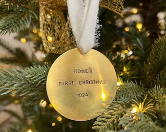 以下が含まれることがあります： 「ROME'S FIRST CHRISTMAS 2024」の文字が入った、丸い金色のクリスマスオーナメント。白いリボンで吊り下げられており、装飾されたクリスマスツリーを背景にしています。