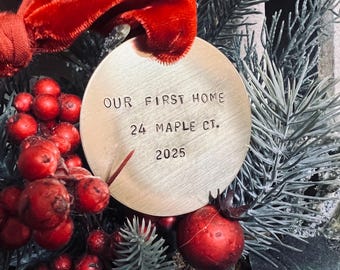 以下が含まれることがあります： 「OUR FIRST HOME 24 MAPLE CT. 2025」の文字が入った丸い金色のオーナメント。赤いベリー、常緑樹の枝、赤いベルベットのリボンに囲まれ、お祝いの雰囲気を醸し出しています。