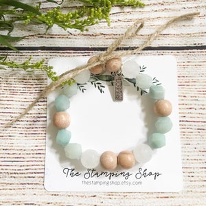 Pode incluir: Um bracelete de contas de madeira e azul claro com um amuleto prateado que diz "Love Never Fails". O bracelete está em um cartão branco com o texto "The Stamping Shop" e "thestampingshop.etsy.com".