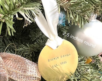 以下が含まれることがあります： 金色の丸いクリスマスオーナメント、直径約6.35cmで、黒字で「RICKEY FAMILY EST. 1995」と書かれています。オーナメントは、緑の針と他のオーナメントが付いたクリスマスツリーの白いリボンから吊り下げられています。