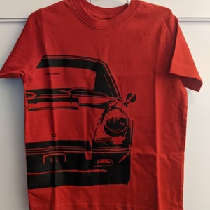 May include: Red t-shirt with a black graphic of a classic car. The car's front end is prominently displayed, featuring headlights and a bumper. The shirt is hanging on a white hanger against a white background.