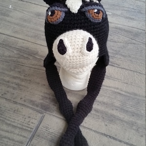 May include: A black and white crocheted horse hat with brown eyes and long black and white tails. The hat is on a white mannequin head.