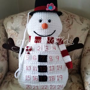 May include: White crocheted snowman advent calendar with a black top hat, red and white striped scarf, and red numbers 1 through 24.