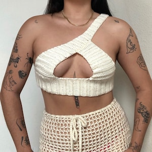 May include: A white crochet crop top with a cut-out design and a criss-cross strap. The top is paired with a white crochet skirt.