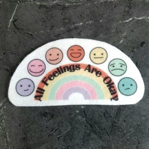 May include: A white felt patch with a rainbow and seven different colored smiley faces. The text "All Feelings Are Okay" is written in black on the patch.