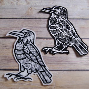 May include: Two black and white embroidered crow patches with intricate designs. The crows are facing different directions and have detailed patterns on their bodies and wings.