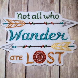 May include: Embroidered patch with the quote "Not all who wander are lost" with a stylized arrow design. The quote is in a teal blue font, the arrows are in shades of brown, orange, and gray, and the background is white.