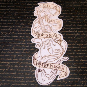 May include: A white embroidered patch with a brown outline of a violin, a pipe, and a banner that reads "Life is full of whimsical happenings".