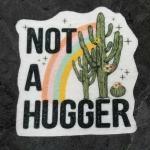 May include: White sticker with a black and white graphic of two cacti with a rainbow behind them. The text "NOT A HUGGER" is printed in black.