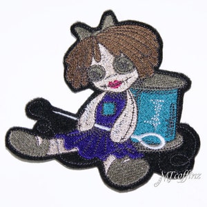 May include: Embroidered patch of a doll with brown hair, wearing a purple dress and sitting on a black and white spool of thread. The doll has a stitched-on smile and large eyes. The patch is surrounded by a black border.
