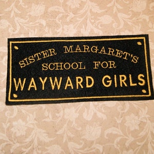 May include: Black sign with gold embroidered text that reads "Sister Margaret's School for Wayward Girls".