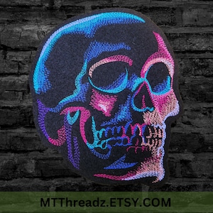 May include: A black felt skull embroidered with blue, pink, and purple thread. The skull is facing right and has a detailed design.
