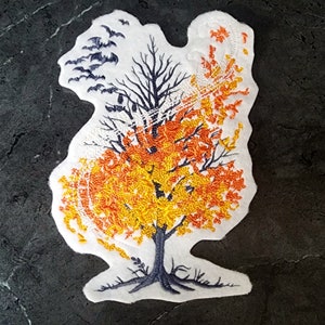 May include: Embroidered patch featuring a tree with autumn leaves in shades of orange and yellow. The tree transitions to bare black branches and a flock of bats. The patch has a white border and is likely for clothing or accessories.