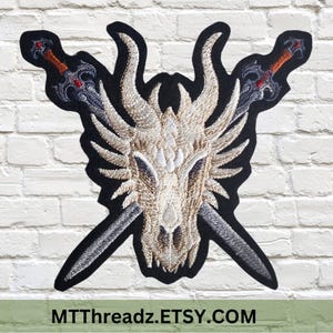 May include: Embroidered patch featuring a white dragon skull with crossed swords. The swords have red and silver accents. The patch is on a black background.