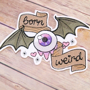 Peut inclure: Patch brodée thermocollante avec un œil rose et violet, des ailes de chauve-souris et deux bannières qui disent "born weird".