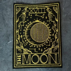 May include: A black felt patch with gold embroidery depicting the The Moon tarot card. The card features a crescent moon, stars, and clouds. The text "The Moon" and the Roman numeral "XVIII" are also embroidered on the patch.