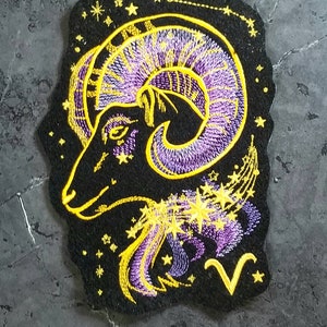 May include: Embroidered patch featuring the Aries zodiac sign. The design showcases a ram's head in purple and yellow thread on a black background. Stars and the Aries symbol are also included.