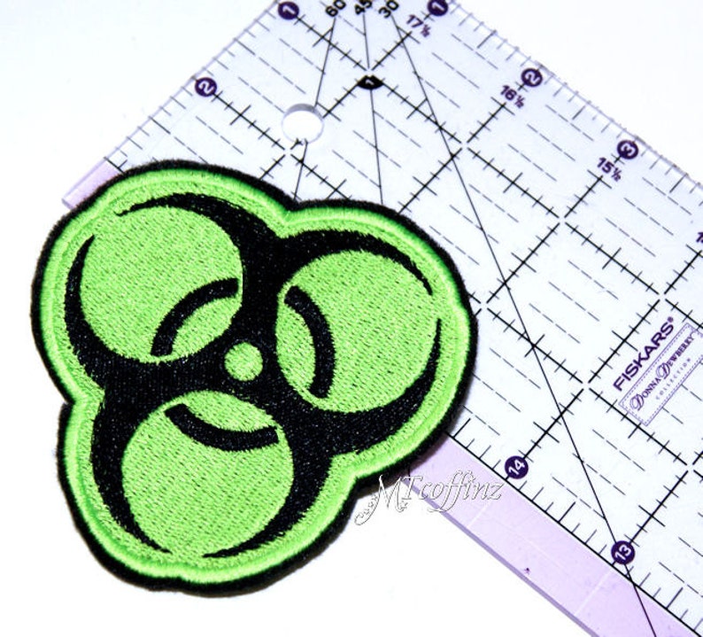 May include: Green and black biohazard symbol embroidered patch. The patch is approximately 5 cm in diameter.