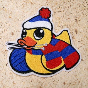 May include: Embroidered patch featuring a yellow duck wearing a blue and white knit cap and a red and blue striped scarf. The duck is holding knitting needles and a ball of blue yarn. The patch has a white border.