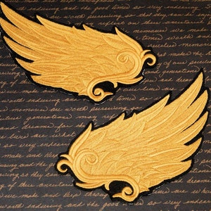 May include: Two gold embroidered wings with black outlines on a dark background. The wings are in a stylized design with a curled tip on each wing.