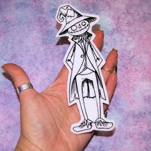 May include: Black and white embroidered iron-on patch of a cartoon scarecrow wearing a hat and a long coat.