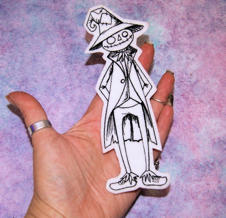 May include: Black and white embroidered iron-on patch of a cartoon scarecrow wearing a hat and a long coat.