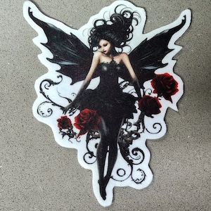 May include: A black fairy with dark hair and wings, holding red roses, against a white background with black floral designs.